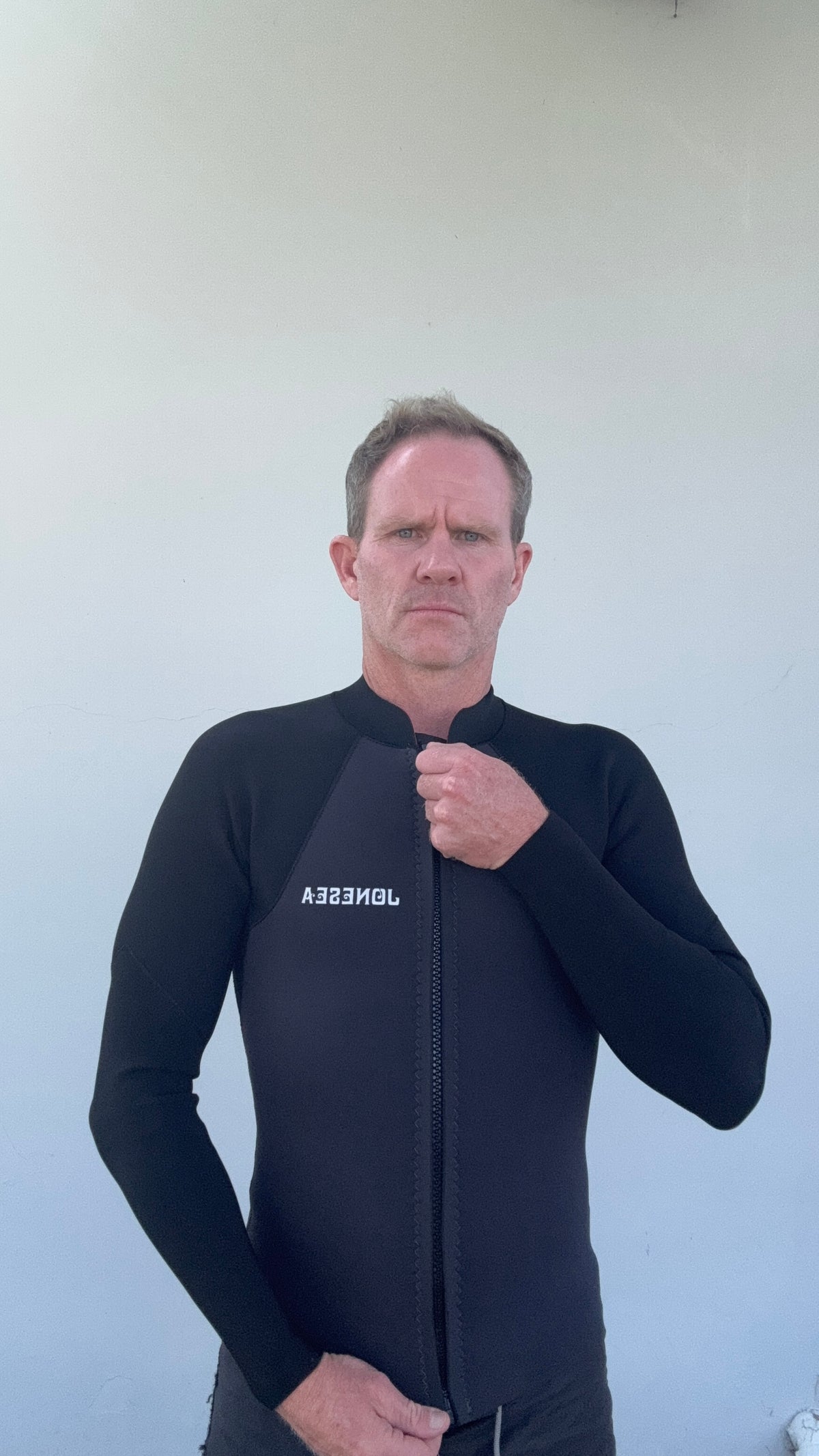 Man wearing the Men’s Graphite Surf Jacket with a front zipper and white branding on the chest, standing against a plain light gray wall.