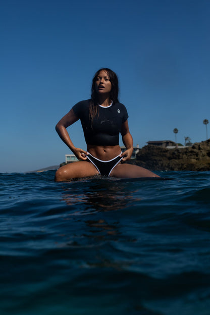 Women wearing black Crop top and a bikini bottom sitting on a surfboard in the ocean. 