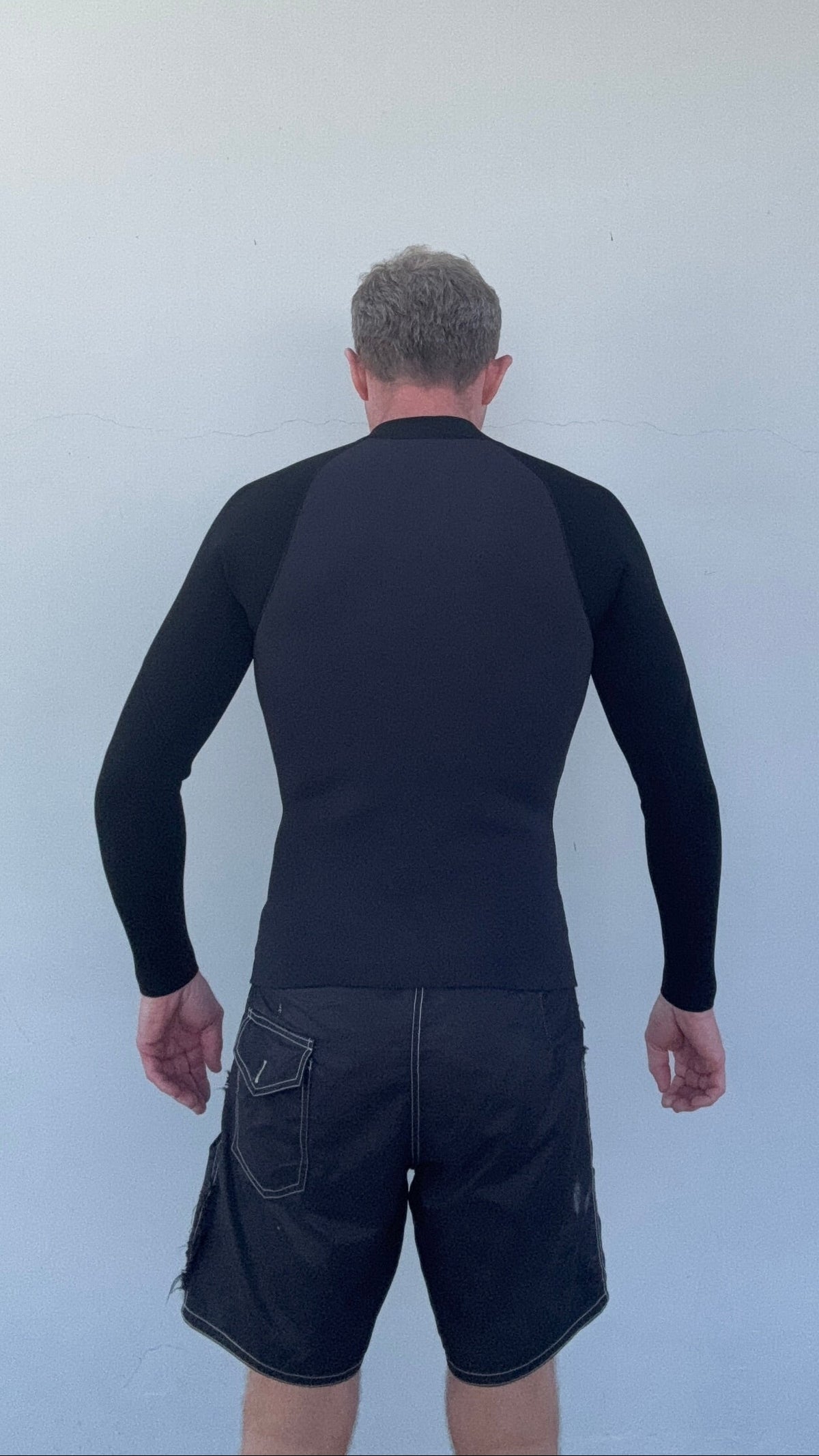 Man wearing the Men’s Graphite Surf Jacket in Graphite, viewed from behind showing the back panel against a plain light gray wall.