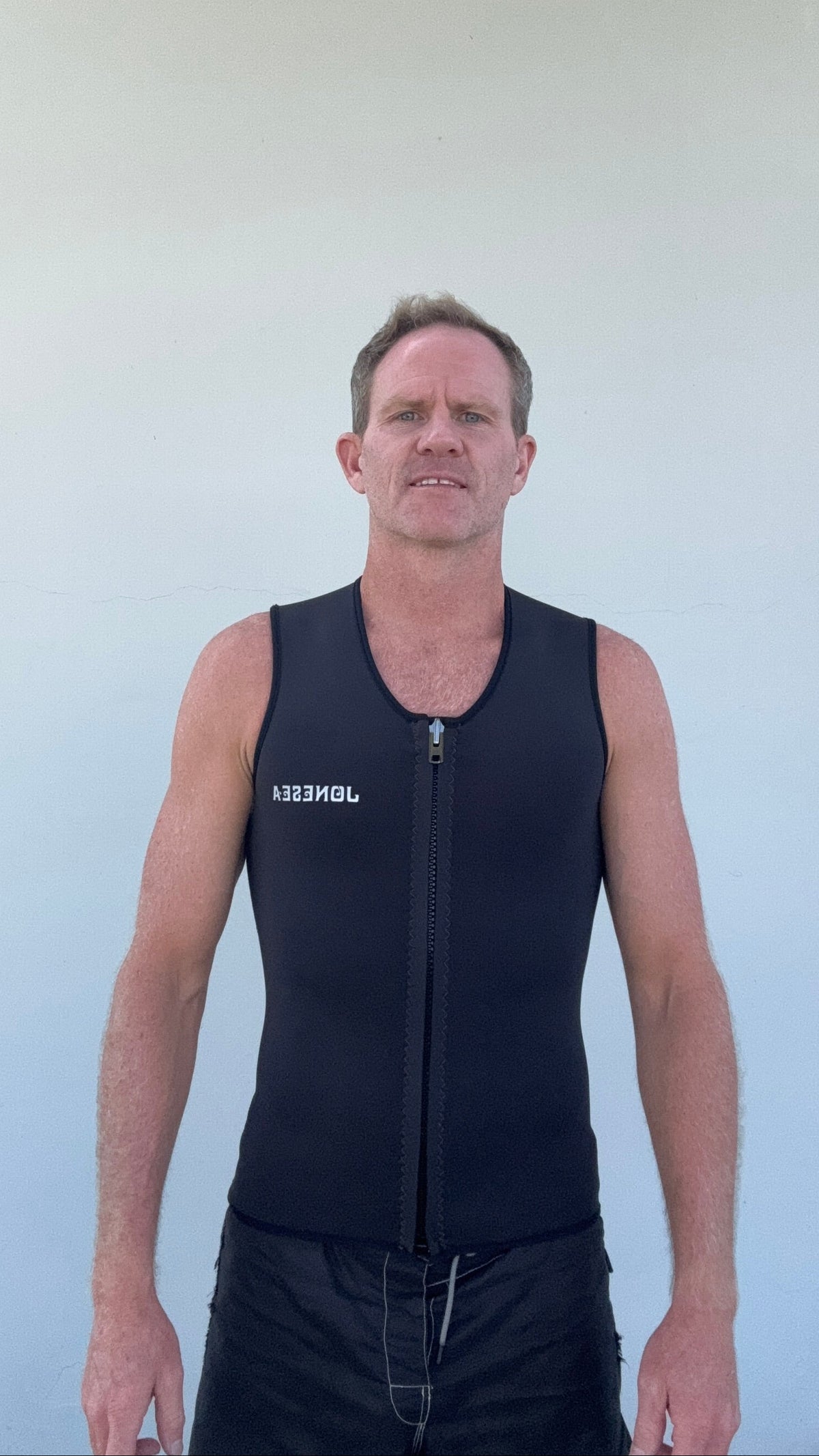 Man wearing the Men's Front Zip Yulex Vest in Graphite with a front zipper, standing against a plain light gray wall.