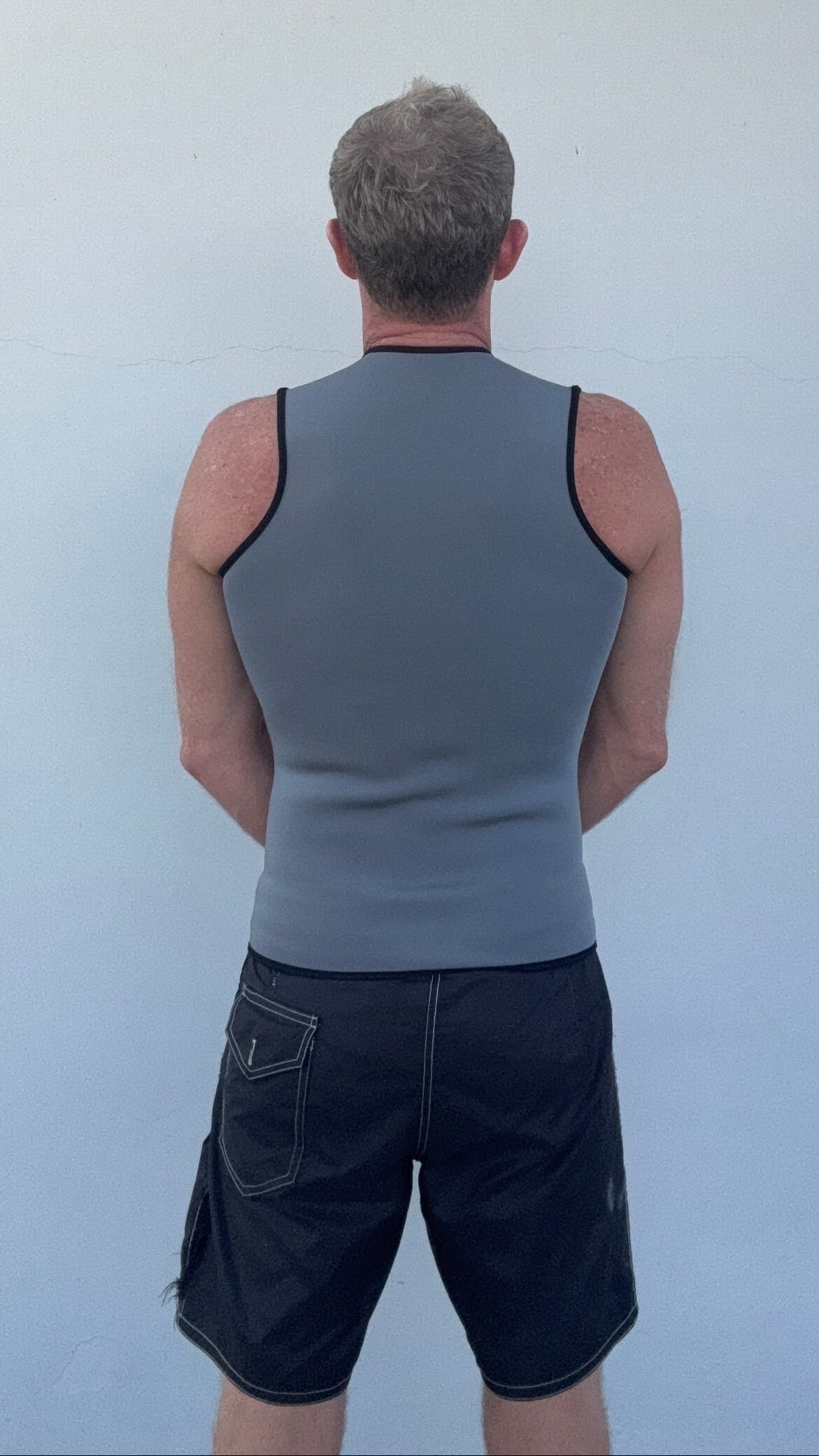 A man viewed from behind wearing a gray neoprene athletic vest against a plain light gray wall.