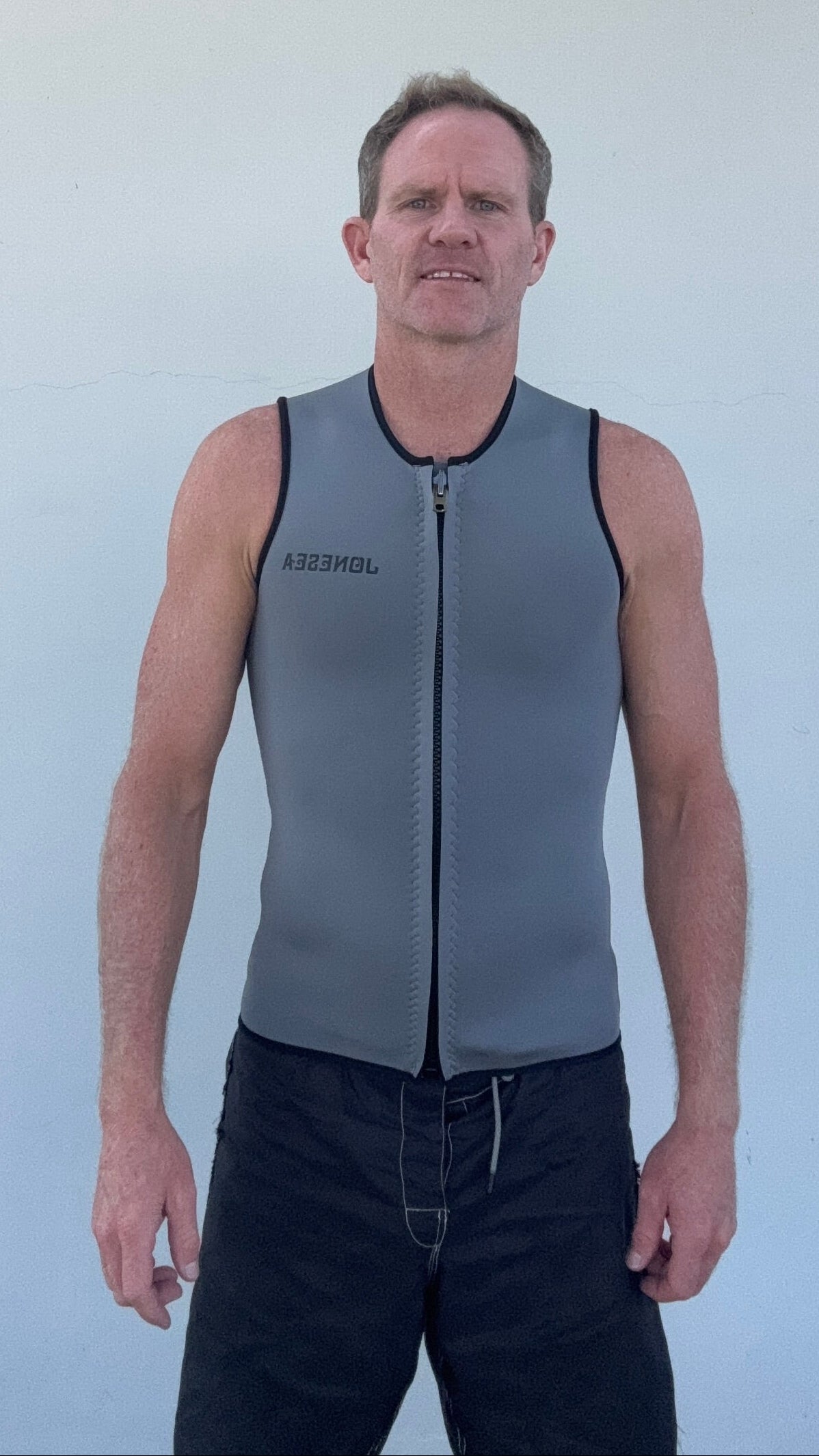 A man wearing a gray neoprene athletic vest with a front zipper and black trim, standing against a plain white wall.